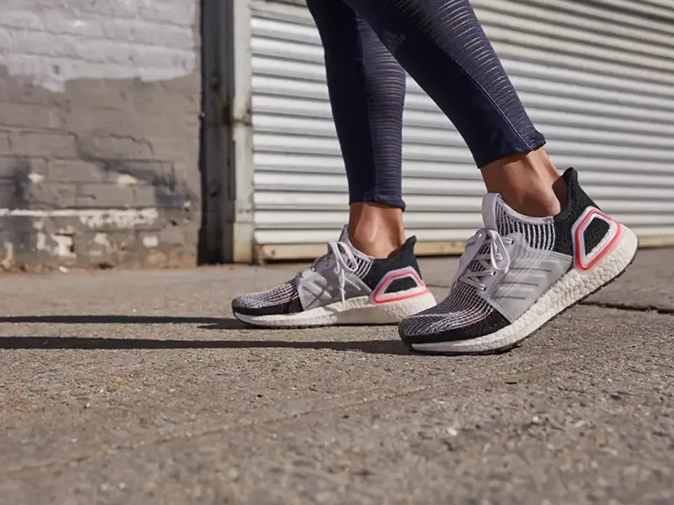 A runner mid-stride in grey and white Adidas Ultraboost 19, shot low against a sunlit roller shutter