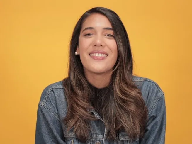 A woman in a denim jacket smiling to camera against a warm yellow backdrop