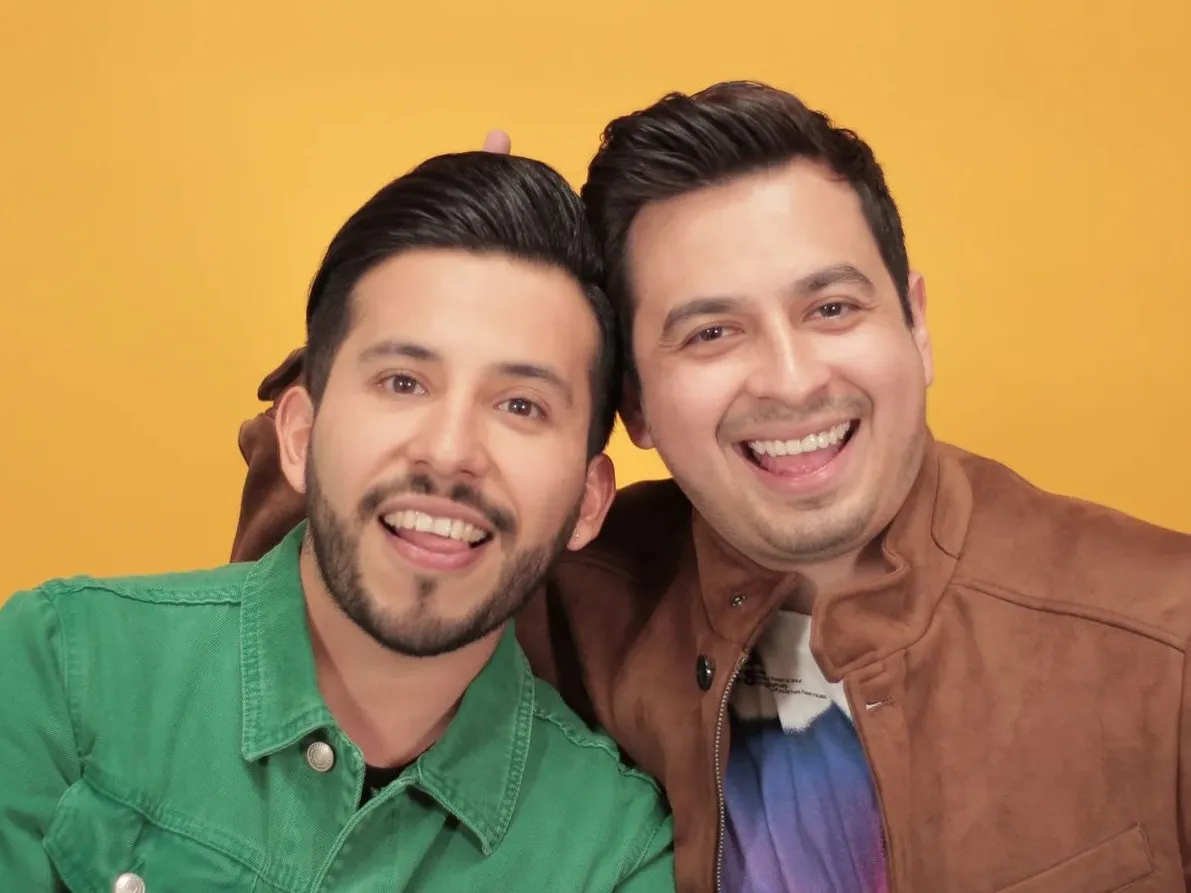 Two men in green and tan jackets leaning head to head, laughing, against a yellow backdrop