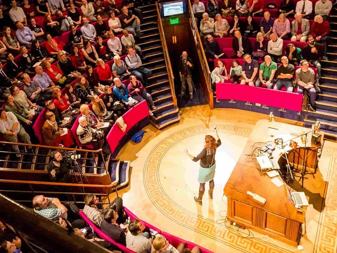 A speaker mid-lecture on the floor of the Ri's Faraday Theatre, ringed by a full house in red tiered seating